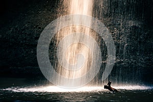 Splashing waterfall with bright sunlight drop on pond with dark cave