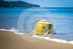 A splashing wave crashes into a pumpkin shaped coconut for Halloween. Green coconut on a tropical beach with a scary face carved