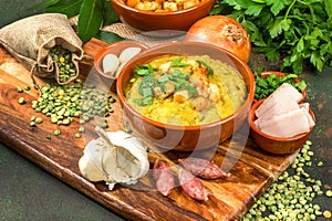 Split pea soup in rustic bowl decorated with fresh green leaves, garlic, onion and slices of lard