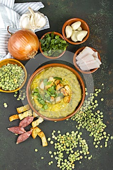 Split pea soup in rustic bowl decorated with fresh green leaves, garlic, onion and slices of lard