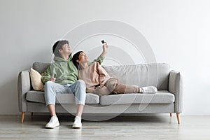 Split system. Happy young Asian couple with remote relaxing under air conditioner, sitting on couch at home, mockup