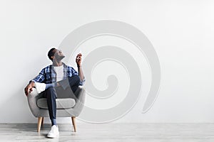 Split system. Young black man with remote turning on air conditioner, sitting in armchair against white wall, mockup