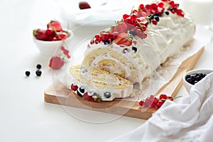 Sponge biscuit cake roll filling whipped cream and berries decorated strawberry, blueberry and red currants on white wooden