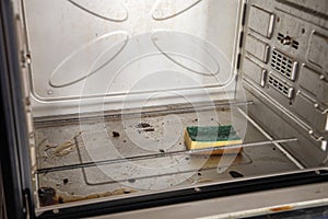 sponge with cleaning solution lies in dirty oven with burnt grease stains on the internal surfaces, freestanding oven