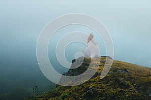 A spooky, blurred, ghostly woman with a white dress, standing on a cliff face looking out on a moody foggy day