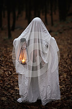 Spooky ghost holding glowing lantern in moody dark autumn forest. Person dressed in white sheet as ghost with light in evening