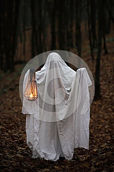 Spooky ghost holding glowing lantern in moody dark autumn forest. Person dressed in white sheet as ghost with light in evening