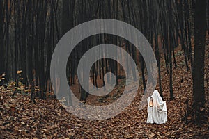 Spooky ghost holding glowing lantern in moody dark autumn forest. Person dressed in white sheet as ghost with light in evening