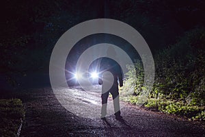 A spooky hooded figure, silhouetted against a cars headlights. On a rainy country road at night