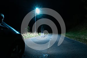 A spooky hooded figure standing below a street light in the countryside. Lit up by car headlights at night