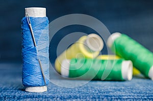 Spool of blue thread with a needle on the background of spools of green and yellow thread on a denim