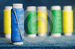 Spool of blue thread with a needle on the background of spools of green and yellow threads on a denim fabric