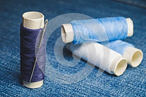 A spool of blue threads with a sewing needle on the background of spools of blue and white threads on a denim fabric