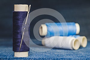 A spool of blue threads with a sewing needle on the background of spools of blue and white threads on a denim fabric