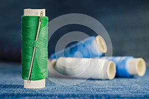 Spool of green thread with a needle on the background of spools of blue and white thread on a denim