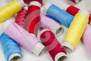 Spools of pink, yellow, blue and red threads and sewing needle on white cotton cloth