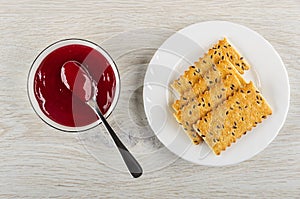 Spoon in bowl with cherry jam, crackers with flax seeds in plate on wooden table. Top view