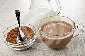 Spoon in bowl of cocoa powder with sugar, pitcher with milk, cup of cocoa on wooden table