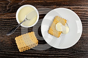 Spoon in bowl with condensed milk, stack of cookies, cookie poured milk in saucer on wooden table. Top view