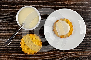 Spoon in bowl with condensed milk, waffle cookies on table, cookie poured condensed milk in saucer. Top view