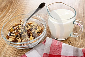 Spoon in bowl of granola with banana and chocolate, pitcher with yogurt, napkin on wooden table