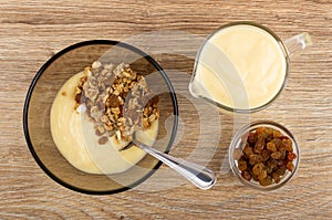 Spoon in bowl with muesli, yogurt, jug of yogurt, bowl with raisin on table. Top view