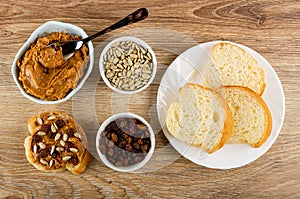 Spoon in bowl with peanut butter, sandwich with peanut pasta, bowls with sunflower seeds and raisin, bread in plate on wooden