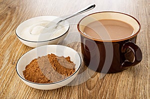 Spoon in bowl with sugar, bowl with cacao powder, cup of cocoa with milk on table