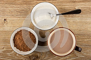 Spoon in bowl with sugar, bowl with cacao powder, cup of cocoa with milk on table. Top view
