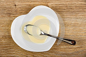 Spoon with condensed milk in saucer in heart shape on wooden table. Top view