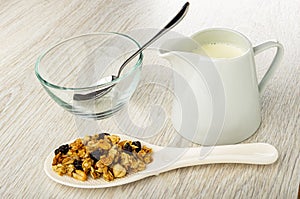 Spoon in empty bowl, jug with yogurt, granola with peanut, hazelnut and raisin in white spoon on wooden table