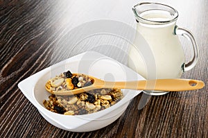 Spoon with granola in bowl with cereal breakfast with peanut and raisin, pitcher with yogurt on wooden table