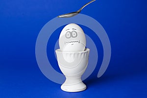 Spoon over egg with drawn frightened face among others in cup on blue background