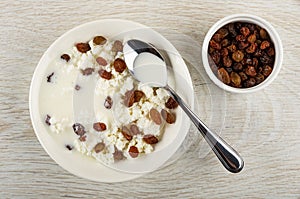 Spoon on plate with cottage cheese, raisin, yogurt, bowl with dried grape on table. Top view