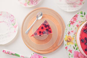 Spoon and red jelly piece of cheesecake with currant on a wooden board. Dish with cake and tea set on a white table