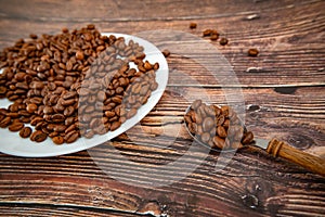 A spoon with roasted coffee lies on a wooden background, next to a white plate with coffee grains