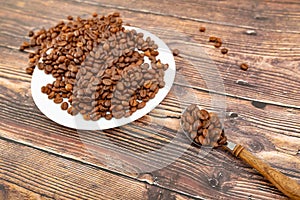 A spoon with roasted coffee lies on a wooden background, next to a white plate with coffee grains