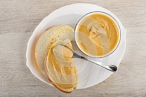 Spoon on sandwich, slice of bread, peanut paste in bowl on dish on wooden table. Top view