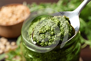 Spoon of tasty arugula pesto near jar with sauce, closeup photo