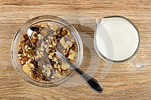 Spoon in transparent bowl of granola with banana and chocolate, pitcher with yogurt on table. Top view