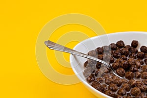 Spoon in White bowl with chocolate corn cereal balls and milk on yellow background, side view. Modern fresh breakfast image