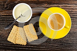 Spoon in white bowl with condensed milk, dry cookies, black coffee in cup on saucer on wooden table. Top view