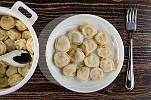 Spoon in plastic pan with boiled dumplings, plate with dumplings, fork on wooden table. Top view