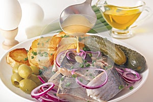 Spoonful of mustard sauce pours over slices of pickled herring with chopped red onions and herbs.