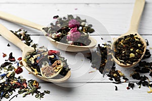 Spoons with dried herbal tea leaves and fruits on white wooden table, closeup