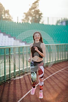 Sport. Athletic young brunette woman in pink sneakers, leggings and top run on running track stadium at sunset. her hair is