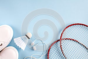 Sport flat lay with white shuttlecock, two rackets for playing badminton, white sneakers and white headphones on a blue background