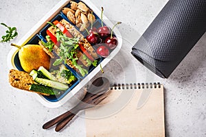 Sport food concept. Lunch box with heathy food, notebook and mat on a gray background