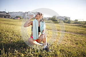 Sport girl drinking water after sport. girl sitting on the grass. building on the background