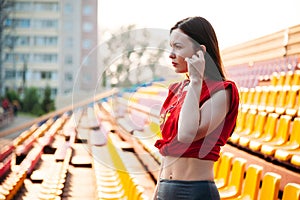 sport girl posing at stadium nad listening to music on headphones. Fitness girl with a sports figure in leggings and red top